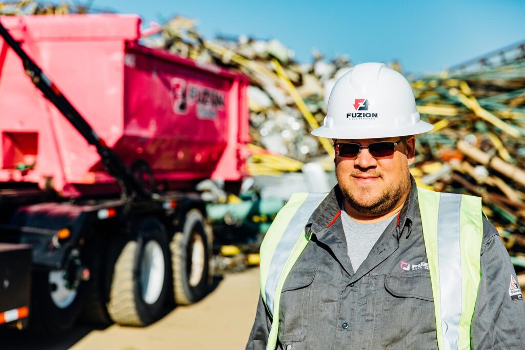A Fuzion worker in a hard hat and safety vest stands in front of a pink dumpster and scrap pile.