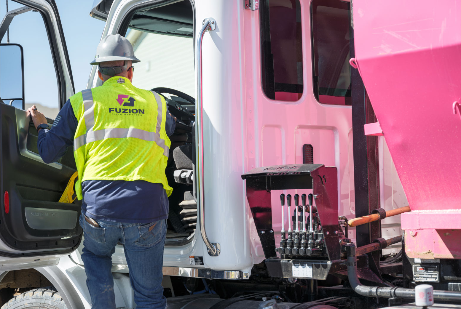 A Fuzion worker operates the controls of a garbage truck beside a pink dumpster.