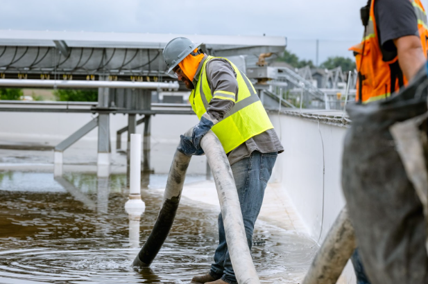 Worker in safety gear using a large hose to manage water at an industrial site.