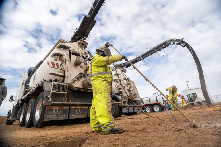Technician in rain gear operating a hydro jetting hose beside a vacuum truck.