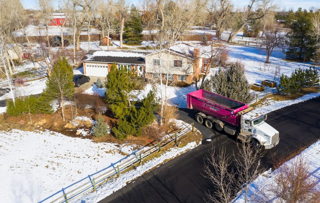 Aerial view of a white truck with a bright pink Fuzion dumpster parked on a residential driveway surrounded by snow.