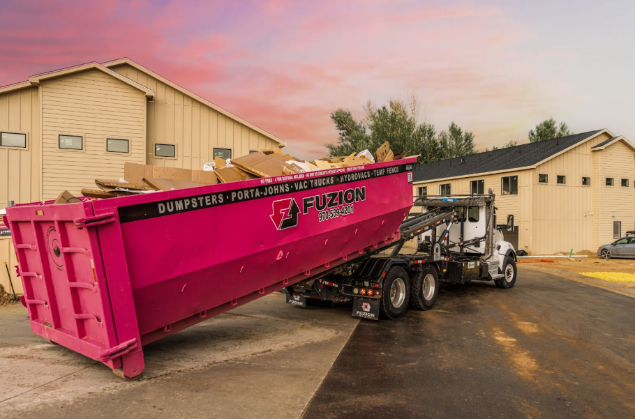 A Fuzion dumpster truck unloads a large pink container at a construction site during sunset.