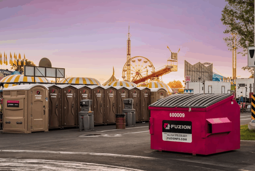 Line of Fuzion portable restrooms and a bright pink dumpster at a fairground during sunset with rides in the background.