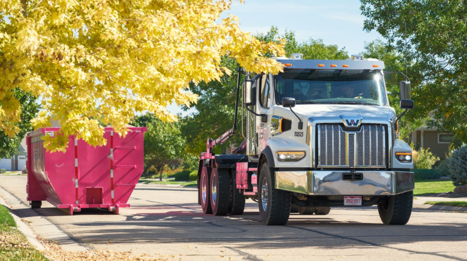 A Fuzion truck approaches a bright pink dumpster on a tree-lined residential street.