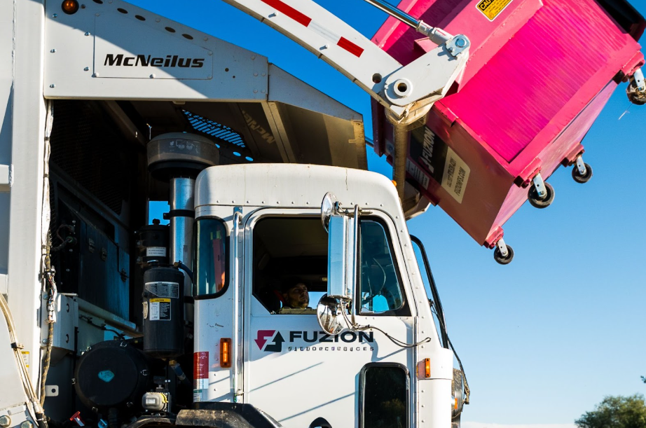 Fuzion Waste truck lifts a bright pink dumpster against a clear blue sky.