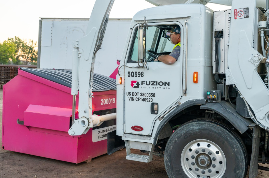 Fuzion Field Services driver operates a truck to lift a pink dumpster.