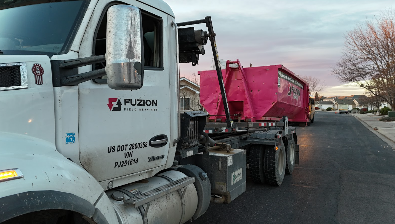 A Fuzion truck hauls a pink container through a quiet neighborhood, showing a dumpster rental for home projects.