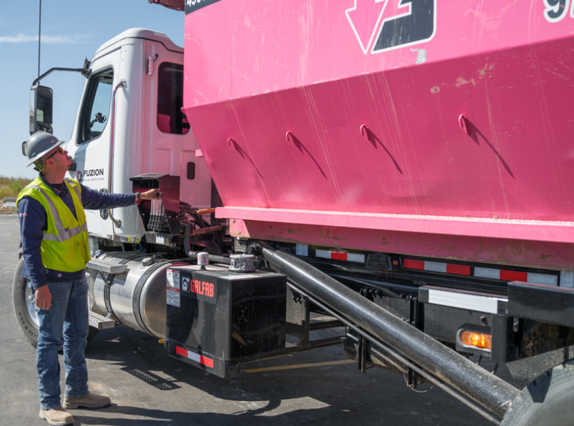 Worker in safety vest and hard hat inspecting a pink roll-off dumpster attached to a white truck.