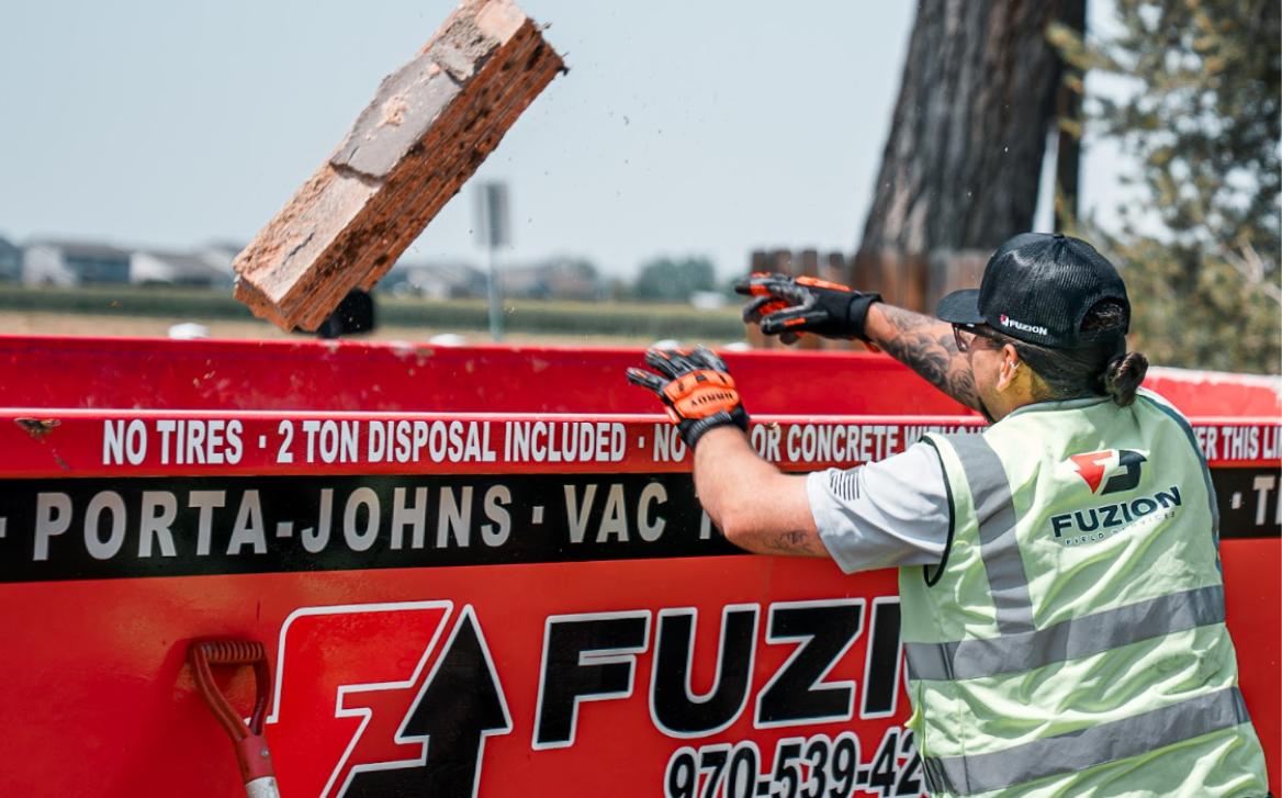 Worker in a Fuzion Field Services vest tossing a chunk of brick into a red Fuzion roll-off dumpster.