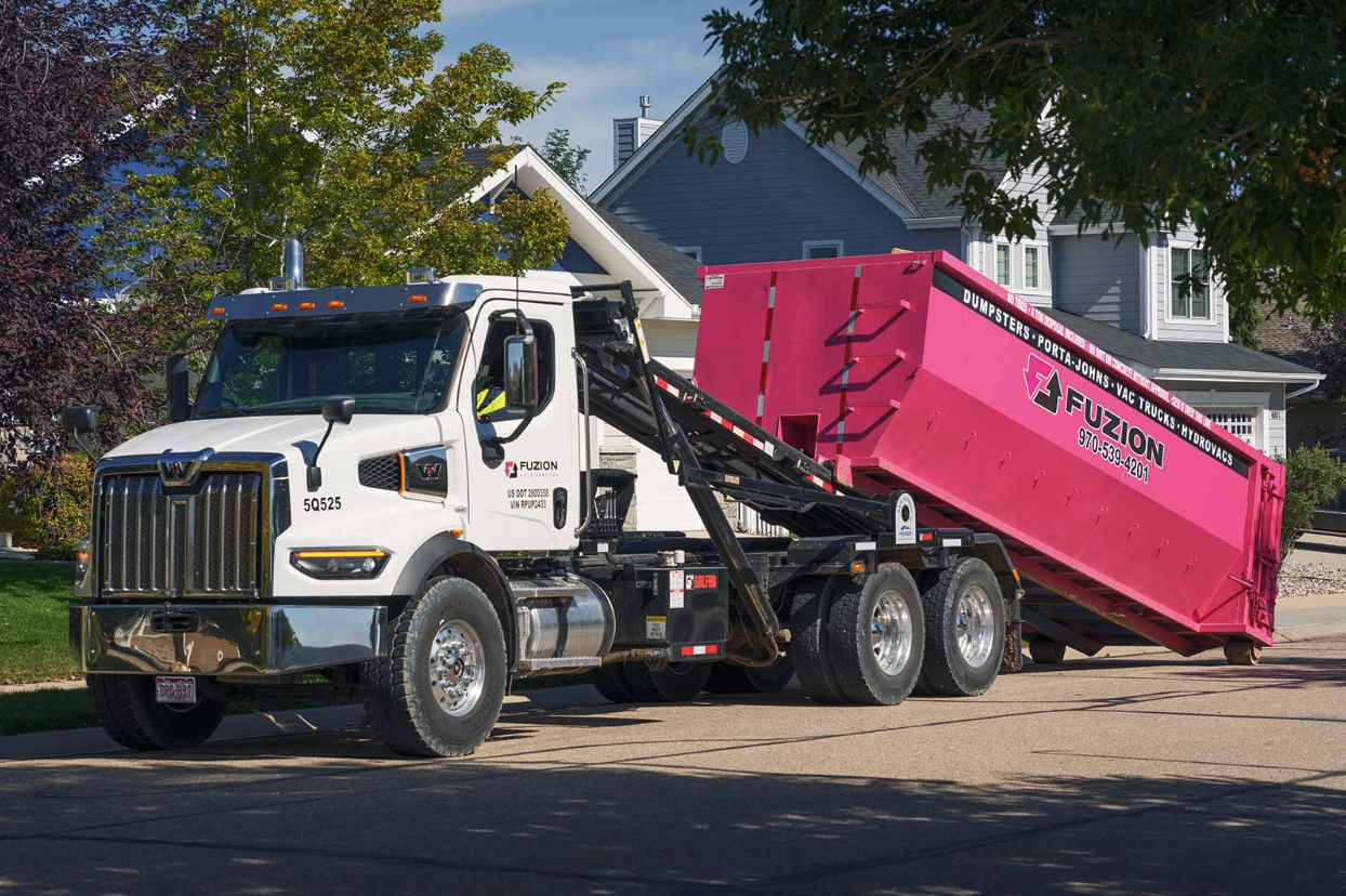 Fuzion roll-off truck delivering a bright pink dumpster to a residential neighborhood construction site.